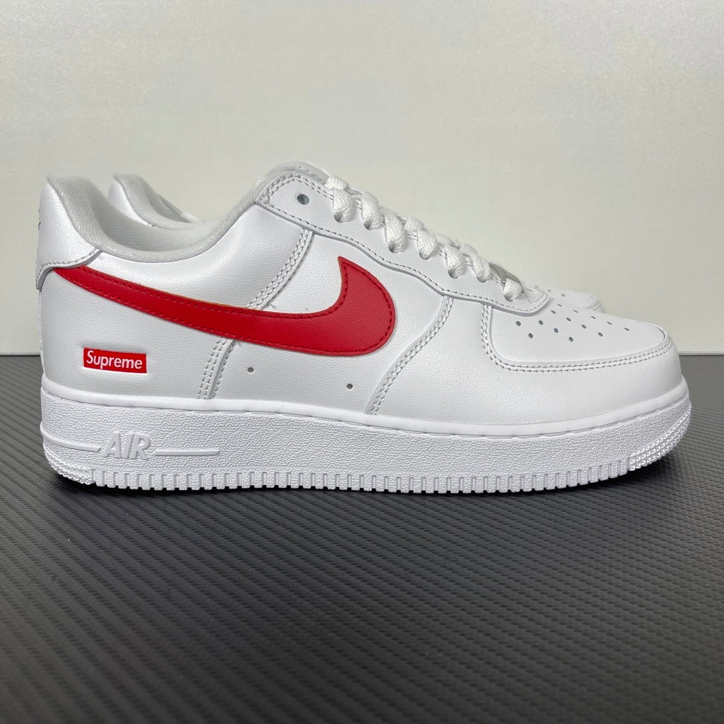 White low-top sneakers with red Nike swoosh and Supreme logo on side, lace-up.