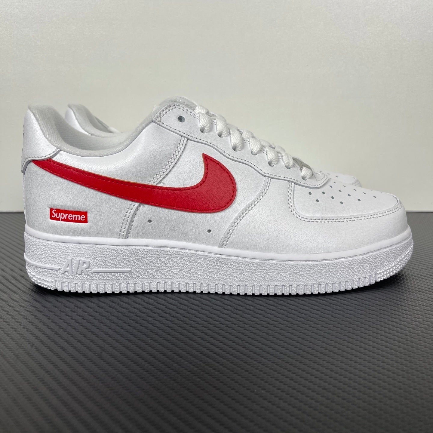 White low-top sneakers with red Nike swoosh and Supreme logo on side, lace-up.