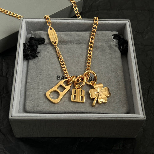 Gold charm necklace with BB pendant, key charm, and clover on a gray jewelry tray