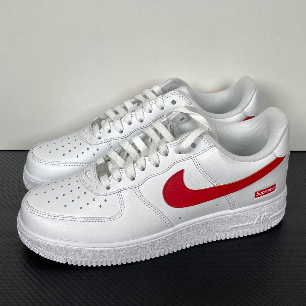 White low-top sneakers with red Nike swoosh and Supreme logo, front side view, lace-up.