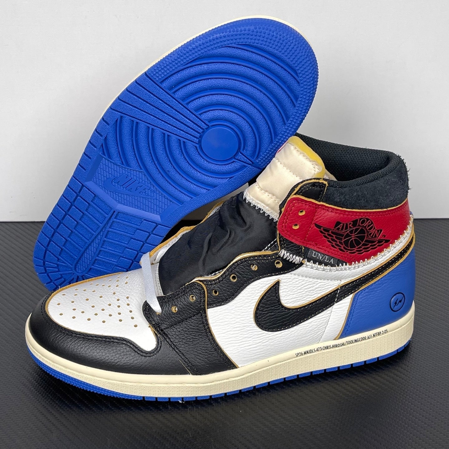 High-top sneaker with blue outsole visible; black and white upper, red ankle patch, gold eyelets