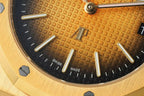 Gold-tone watch dial close-up: black and amber texture, baton markers, date window