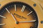 Gold-tone watch dial close-up with black textured pattern, three hands, and logo text