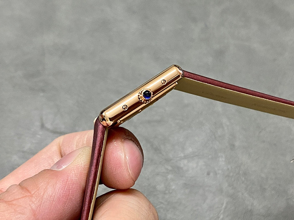 Gold-toned watch case side with crown and blue accent, burgundy leather strap