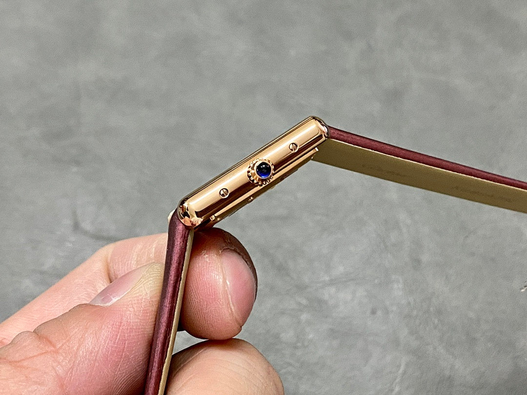 Gold-toned watch case side with crown and blue accent, burgundy leather strap