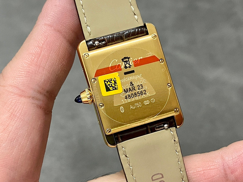 Rectangular watch case with gold-tone back and brown strap; QR-style code and markings visible