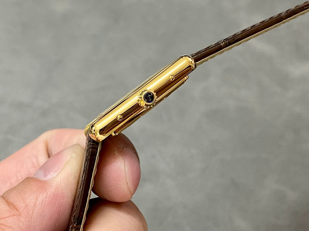 Gold-tone side of a rectangular wristwatch with a round jewel detail and brown leather strap