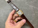 Rectangular wristwatch with Roman numeral dial, gold-tone case, and brown leather strap in hand