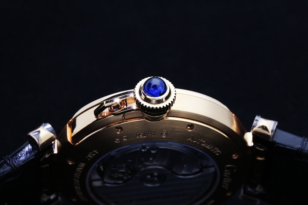 Cartier watch crown with blue gem on gold-tone bezel above dark leather strap