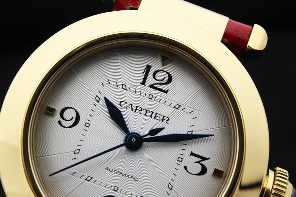 Cartier watch dial close-up with gold bezel, white dial, blue hands, red leather strap