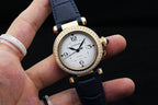 Gold-tone Cartier watch with white dial, diamond bezel, blue leather strap held in hand