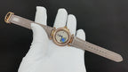 Round-faced wristwatch on a light leather strap with gold-tone case and blue detail