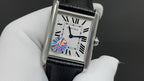 Cartier watch with black leather strap, silver case, white Roman numeral dial and blue gem.