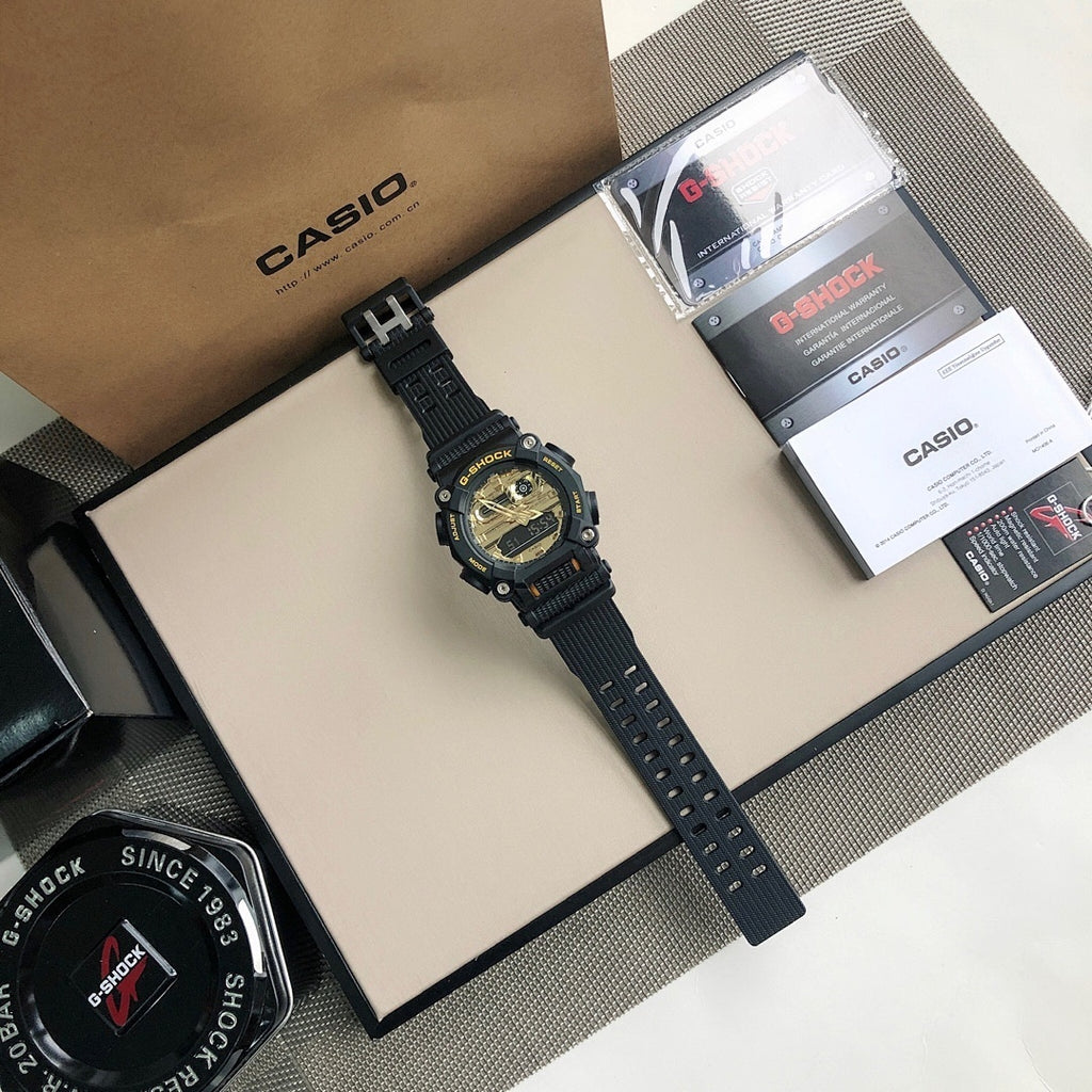 Black G-SHOCK wristwatch on strap with gold dial, shown near accessories