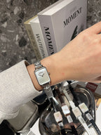 Rectangular wristwatch with silver-tone metal bracelet and white dial held beside boxes