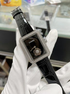 Chanel watch with black leather strap, jeweled rectangular case, dark dial with Chanel logo.