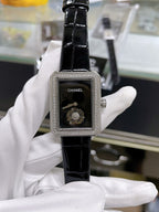Chanel watch with black leather strap, rectangular case, dark dial, hands and Chanel logo.