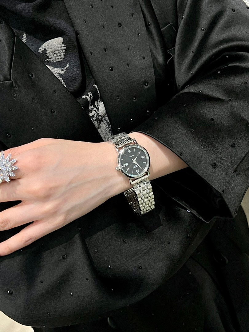 Silver metal bracelet wristwatch with dark dial, worn under a black jacket sleeve.