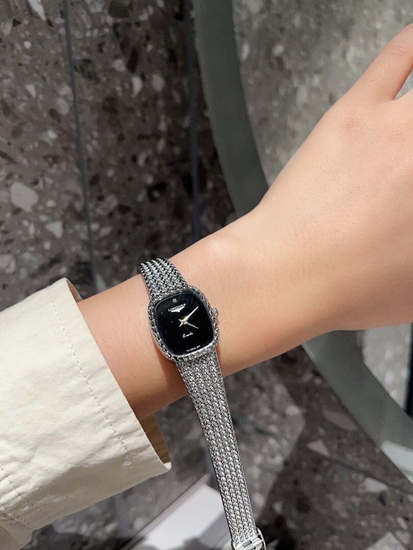 Square wristwatch with black dial and silver mesh bracelet on wrist