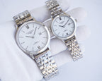 Two-tone wristwatch pair with silver dials, Roman numerals, and metal link bracelets