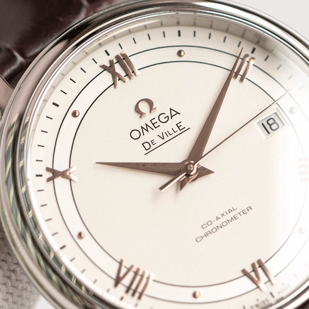 Omega De Ville watch face close-up with white dial, gold markers, hands, date at 18