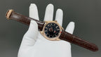 Analog wristwatch with black dial, gold-tone case, and brown leather strap