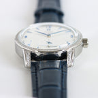 Silver watch with blue leather strap, diamond bezel, white dial and blue accents.