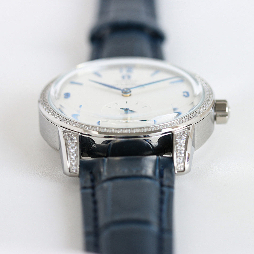 Silver watch with blue leather strap, diamond bezel, white dial and blue accents.