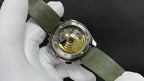 Wristwatch open caseback showing gold-toned movement, green rubber strap on white gloves