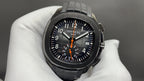 Patek Philippe wristwatch with black dial, orange center hand, and black rubber bracelet