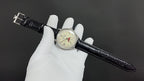 Wristwatch with black leather band extended across gloves; cream dial with Roman numerals
