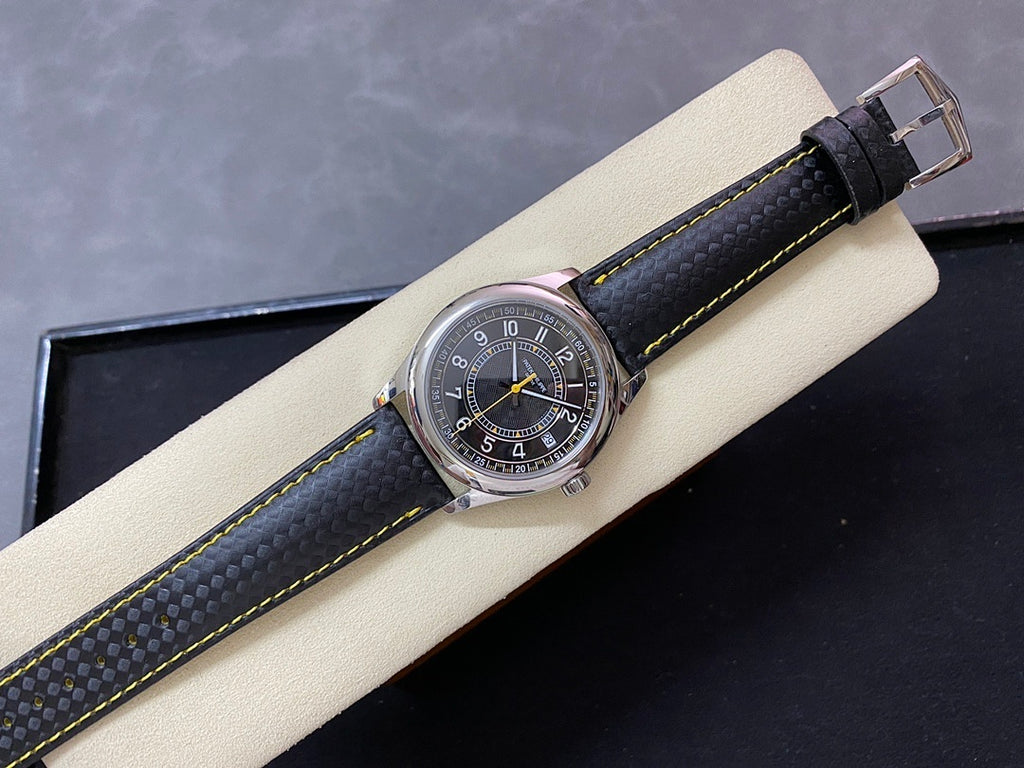 Luxury wristwatch on display pillow with stainless case, dark dial, and black strap with yellow stitching.