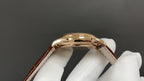 Gold-tone wristwatch case with crown and brown leather strap, angled view