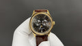 Patek Philippe wristwatch with dark dial, gold bezel, brown leather strap and crown