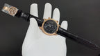 Luxury wristwatch on glove with black dial, gold-tone case, and black leather strap.