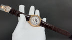 Gold-tone luxury wristwatch on brown leather strap, angled view with white dial