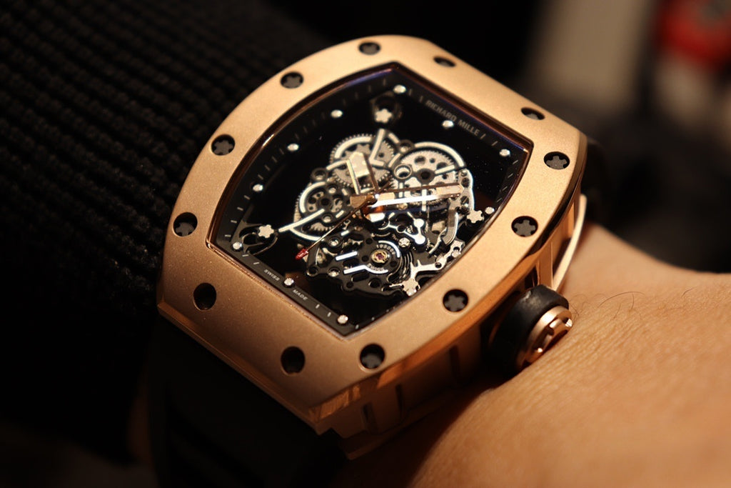 Gold-toned case watch with black rubber strap, tilted view of exposed mechanical movement