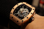 Gold-toned case watch with black rubber strap, tilted view of exposed mechanical movement
