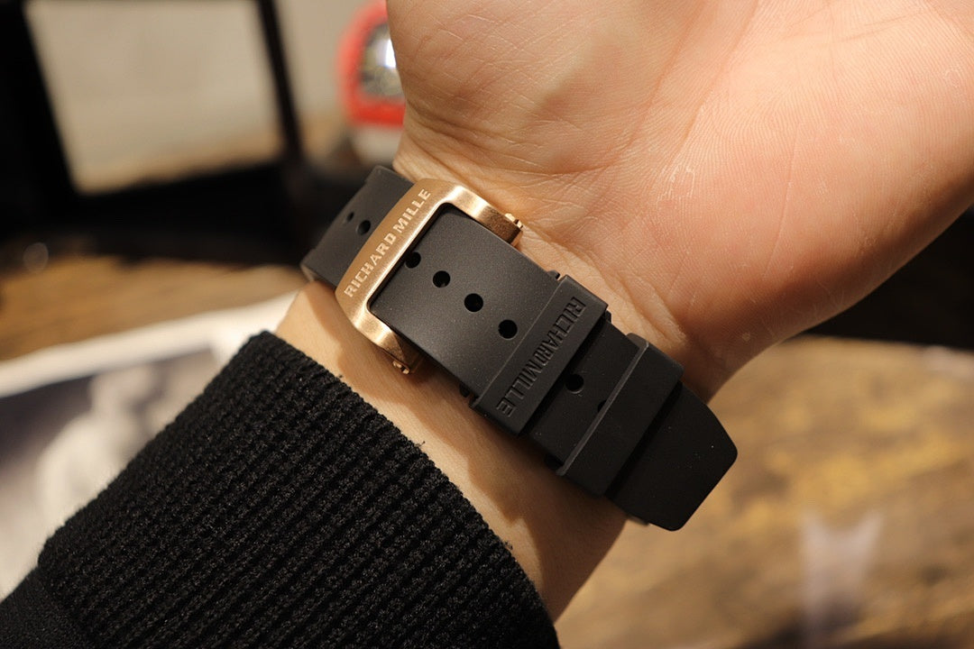 Gold-toned case watch with black rubber strap, clasp side view held in hand