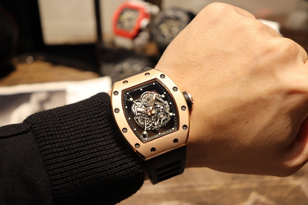 Gold-toned tonneau watch with black rubber strap on wrist, skeleton dial detail