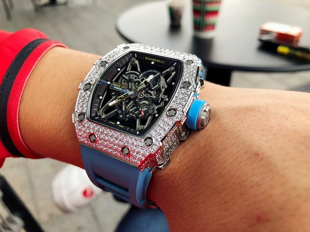 Richard Mille watch on wrist with silver gem-set bezel, blue rubber strap, exposed movement.