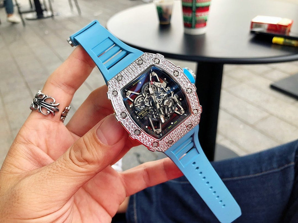 Richard Mille watch held in hand with silver gem-set bezel, blue rubber strap, detailed dial.