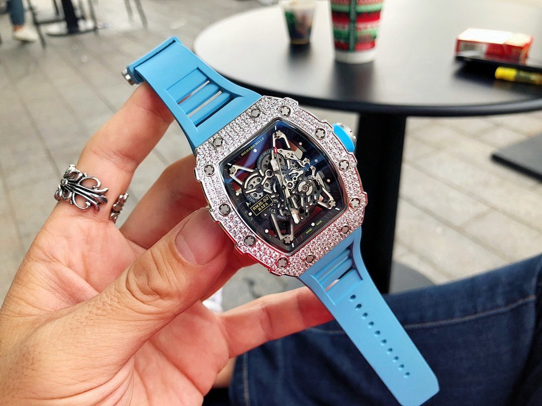 Richard Mille watch held in hand with silver gem-set bezel, blue rubber strap, detailed dial.