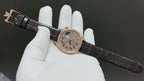 Rose-gold dress watch with black leather strap, crown side view on white gloves.
