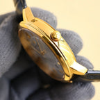 Gold-tone wristwatch side view with textured crown and black leather strap.