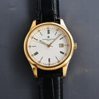 Gold wristwatch with white dial and black leather strap, date window showing 15