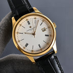 Gold wristwatch front at angle with white dial, black leather strap, date window showing 15