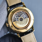 Gold watch with open caseback showing mechanical movement; beige leather strap.