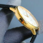 Gold-tone wristwatch case side with crown and curved bezel, on black leather strap.
