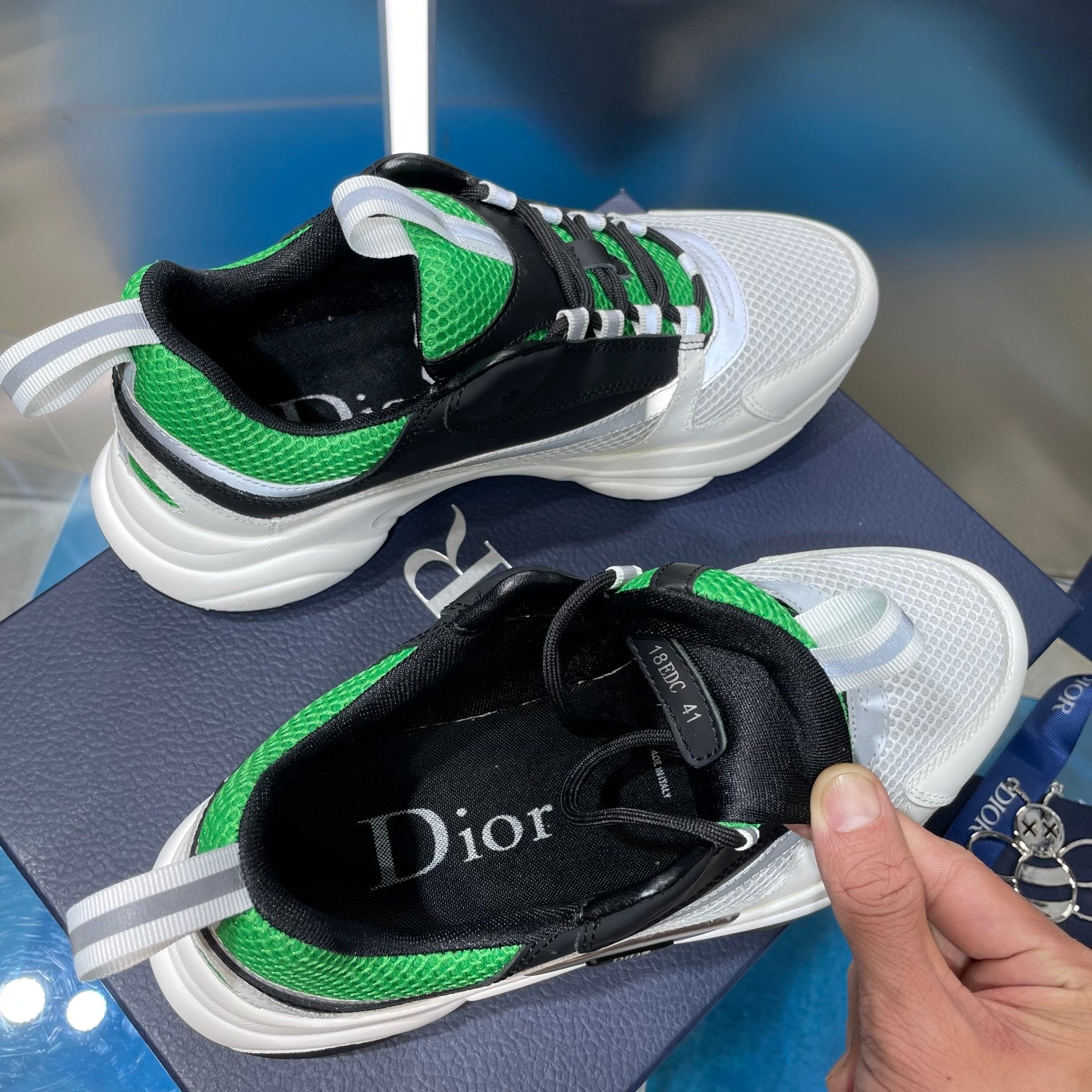 Unisex Dior sneakers close-up, hand holding black tongue; green collar and laces